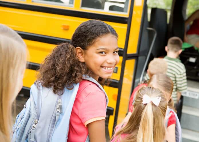 A smiling girl wearing a backpack stands in line with other children as they board a yellow school bus. She looks back over her shoulder at the camera.
