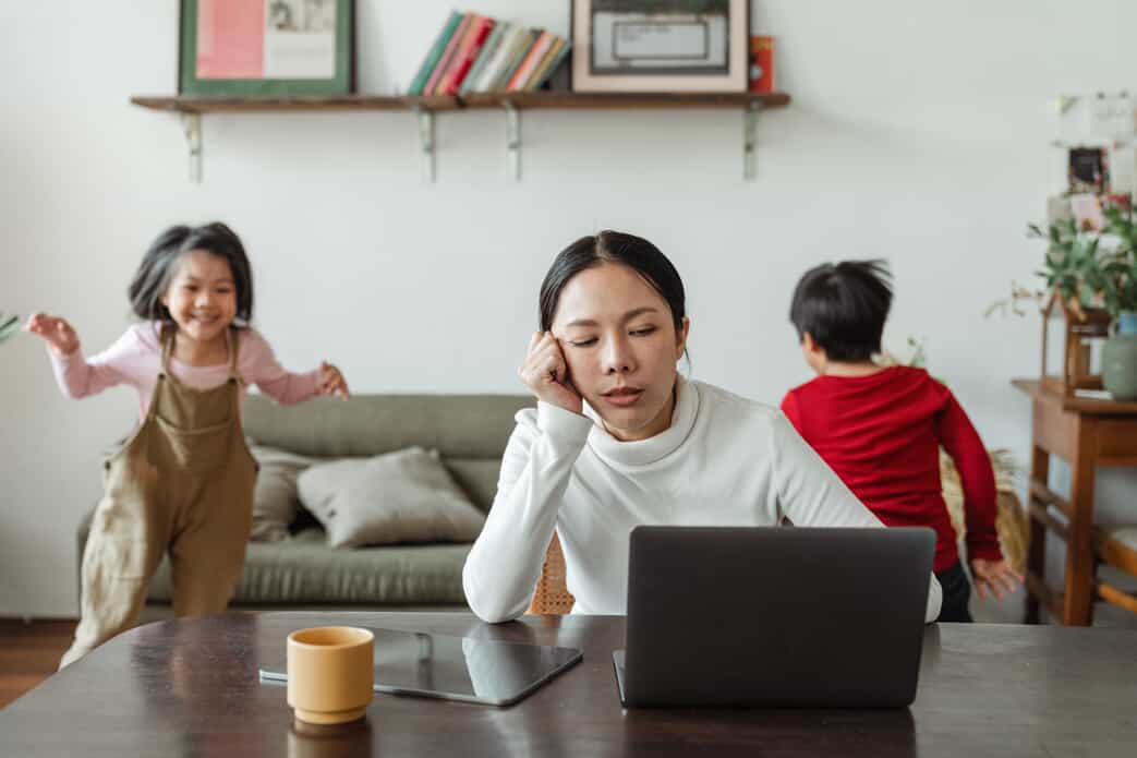 A woman sits at a table with a laptop, looking tired and resting her head on her hand, while two children play energetically in the background. A mug and tablet are on the table.