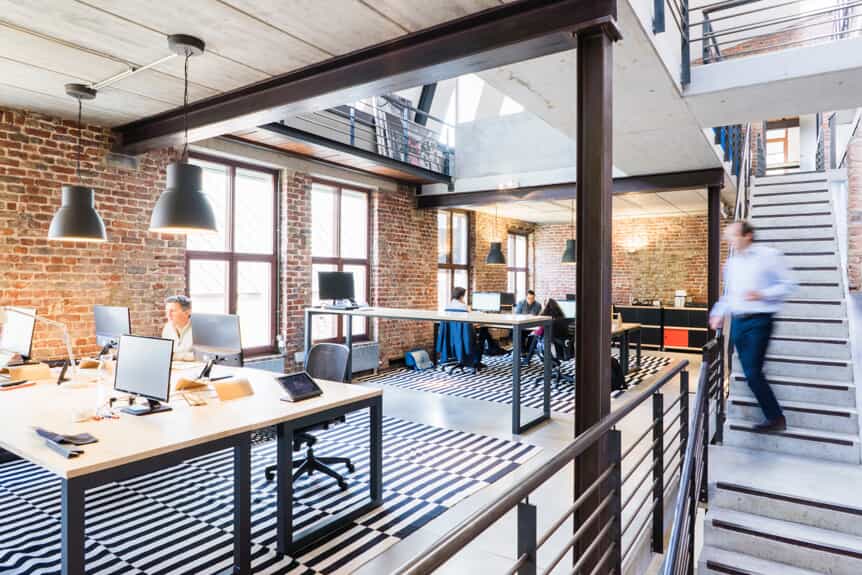 Modern open-plan office with exposed brick walls, large windows, and multiple desks with computers. A person is walking down the stairs, while others work at desks. Black-and-white striped rugs cover the floor.