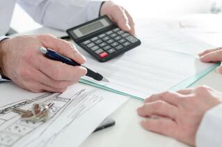 Two people reviewing documents at a desk, one holding a pen and pointing to a form, while the other holds a calculator. Architectural plans and keys are also on the table.
