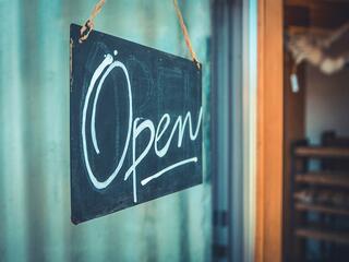 A chalkboard sign with the word Open written in white cursive hangs by two ropes at the entrance of a shop or café. The background is slightly blurred, showing part of the interior.