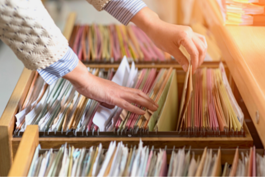 A person’s hands sorting through colorful file folders organized in a wooden filing cabinet drawer, with sunlight illuminating the files.