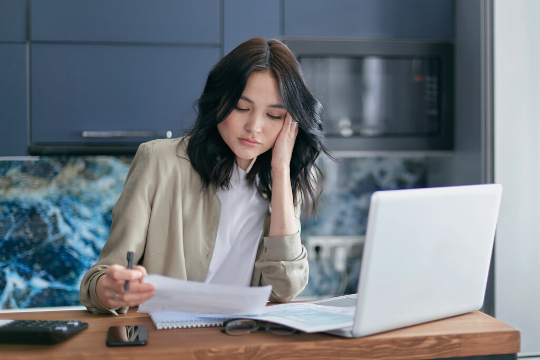 A woman sits at a table with a laptop, holding papers in one hand and a pen in the other, looking focused and slightly tired as she works. There is a calculator and a phone on the table beside her.