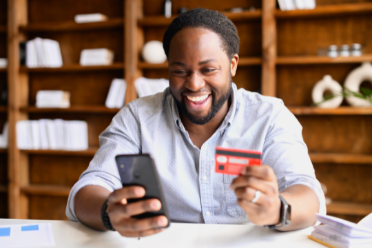 A smiling man sits at a desk holding a smartphone in one hand and a credit card in the other, with shelves of books and decor in the background.