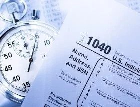 A close-up of a stopwatch next to a U.S. 1040 tax form, emphasizing the urgency of tax filing. Blurred financial documents are visible in the background.