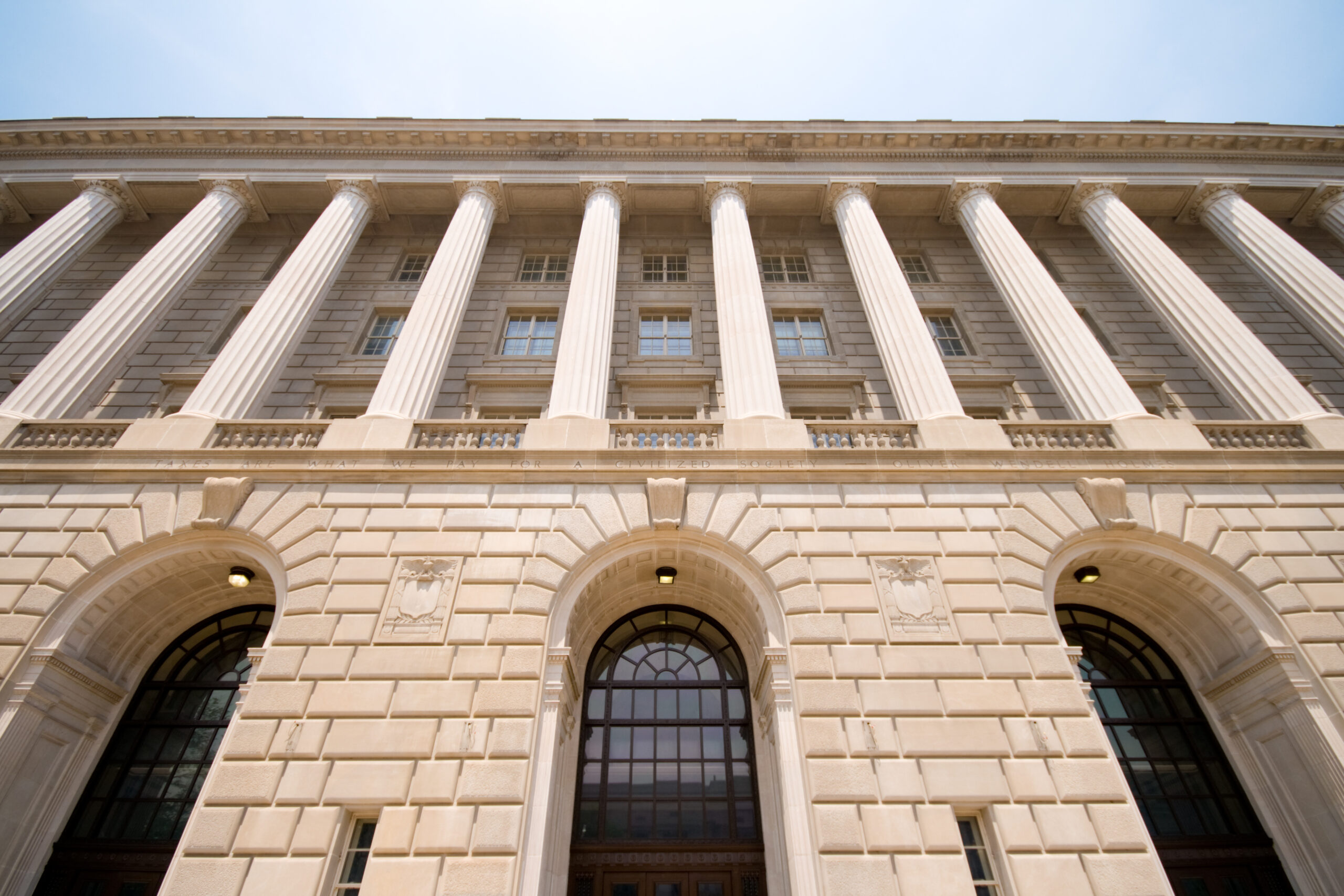 A low angle image of the Internal Revenue Service building in Washington, D.C. It has tan brick with large archways and many columns, stretching up into a blue sky.