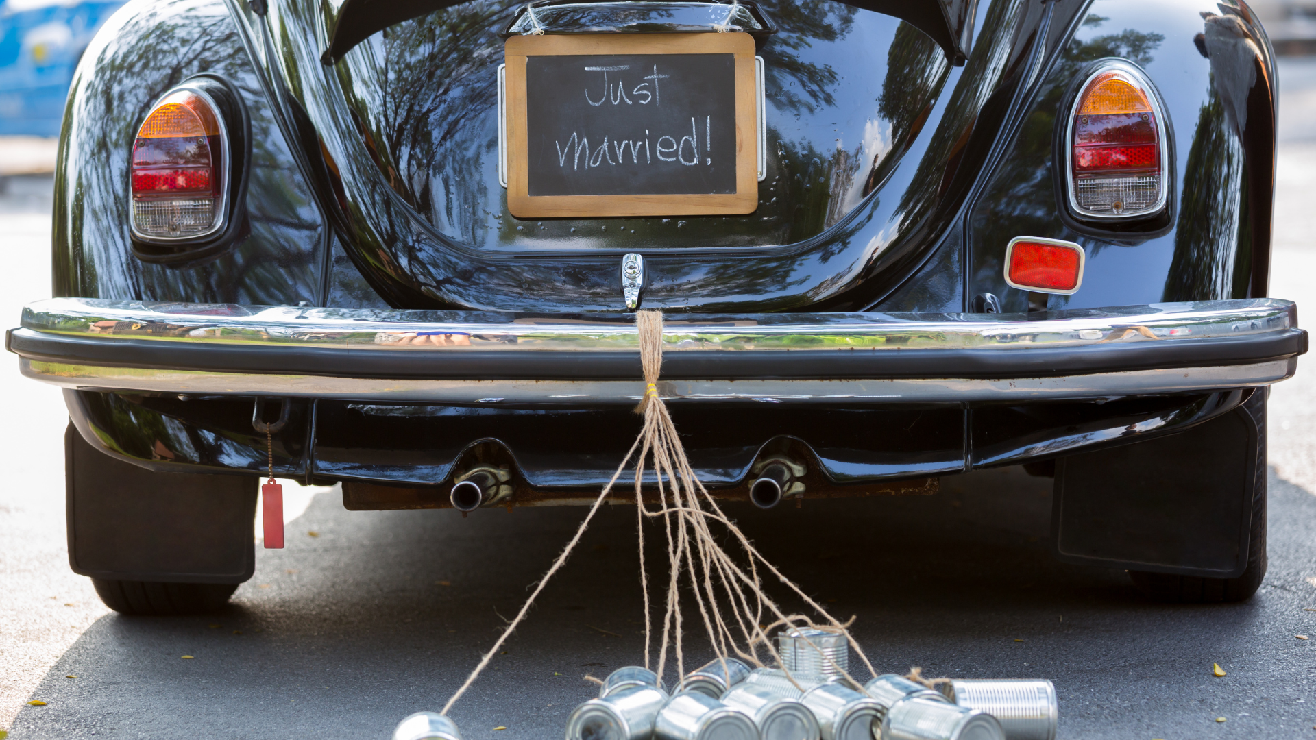A black vintage car is ready to drive away, with a chalkboard "Just Married" sign and a bundle of tin cans affixed to the back