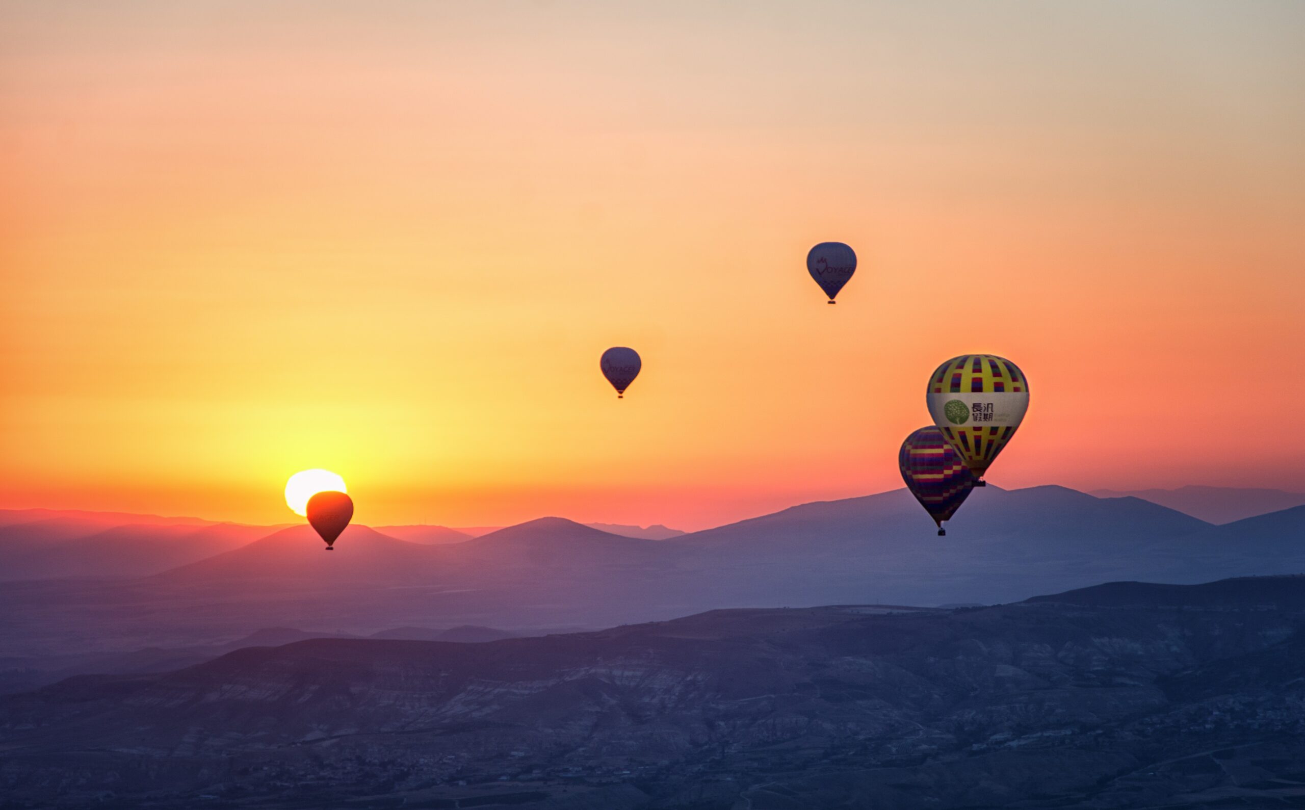 Four hot air balloons float in the sky at sunrise over distant mountains, their gentle ascent echoing inflation prices rising, as the sun peeks above the horizon and soft orange and purple hues fill the sky.