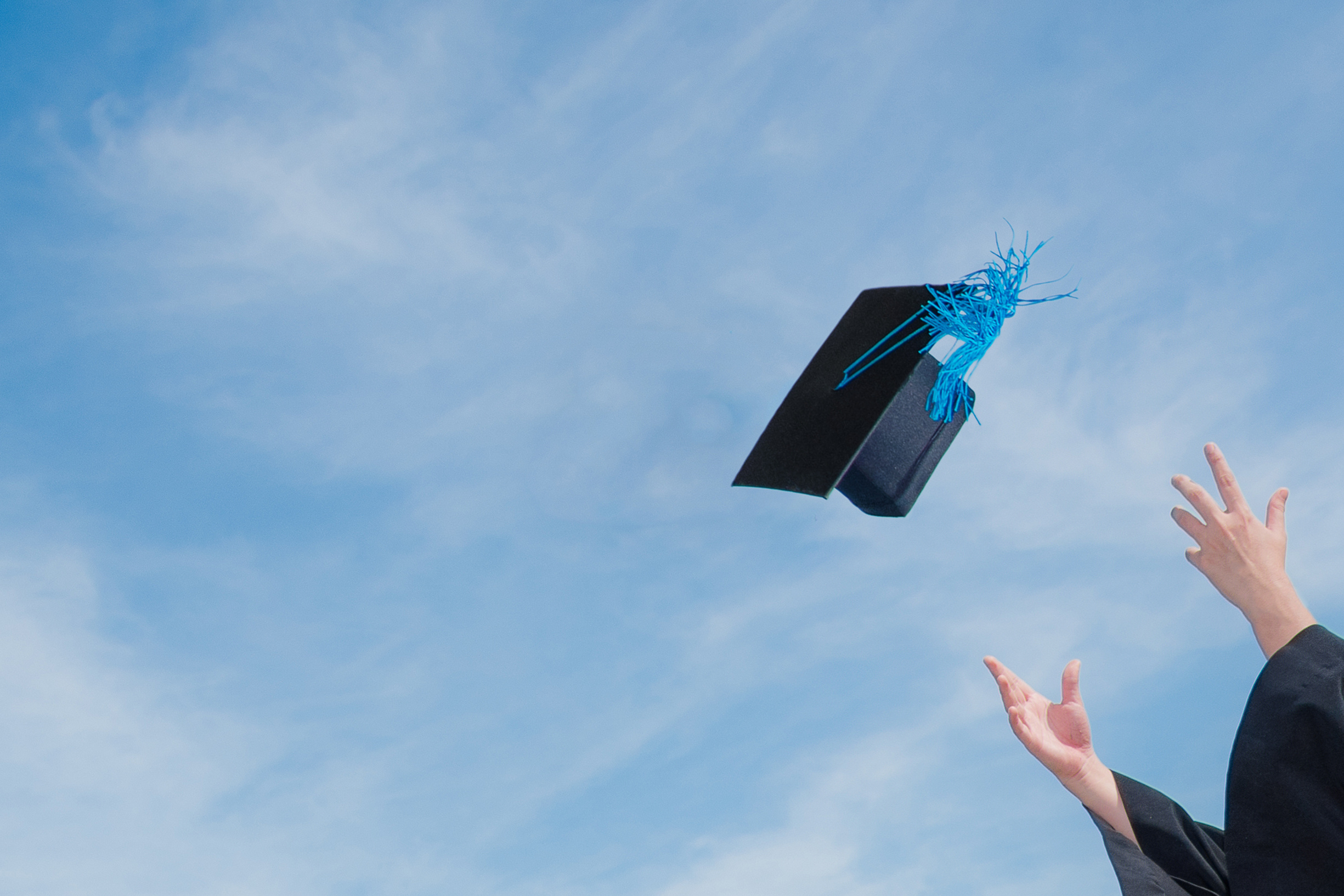 A student tosses a black graduation cap into the sky, which is blue with hazy white clouds