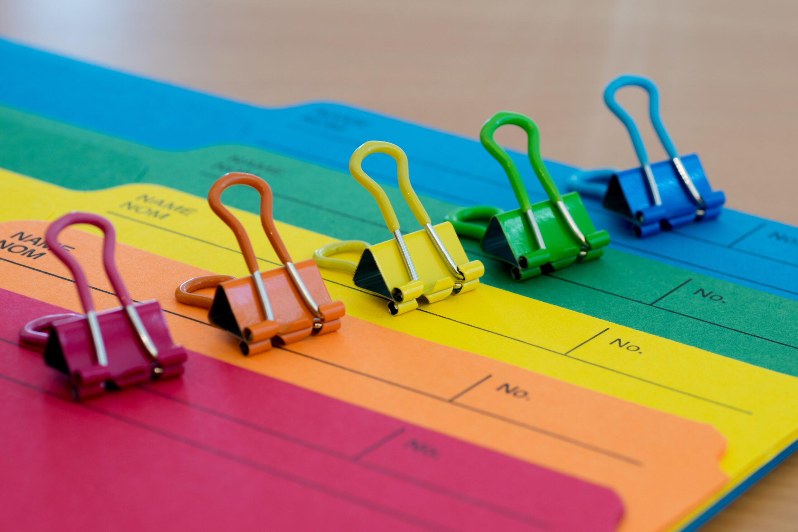 Five brightly-colored file folders and coordinating paper clips are laid out diagonally, forming a rainbow from red, orange, yellow, green and blue