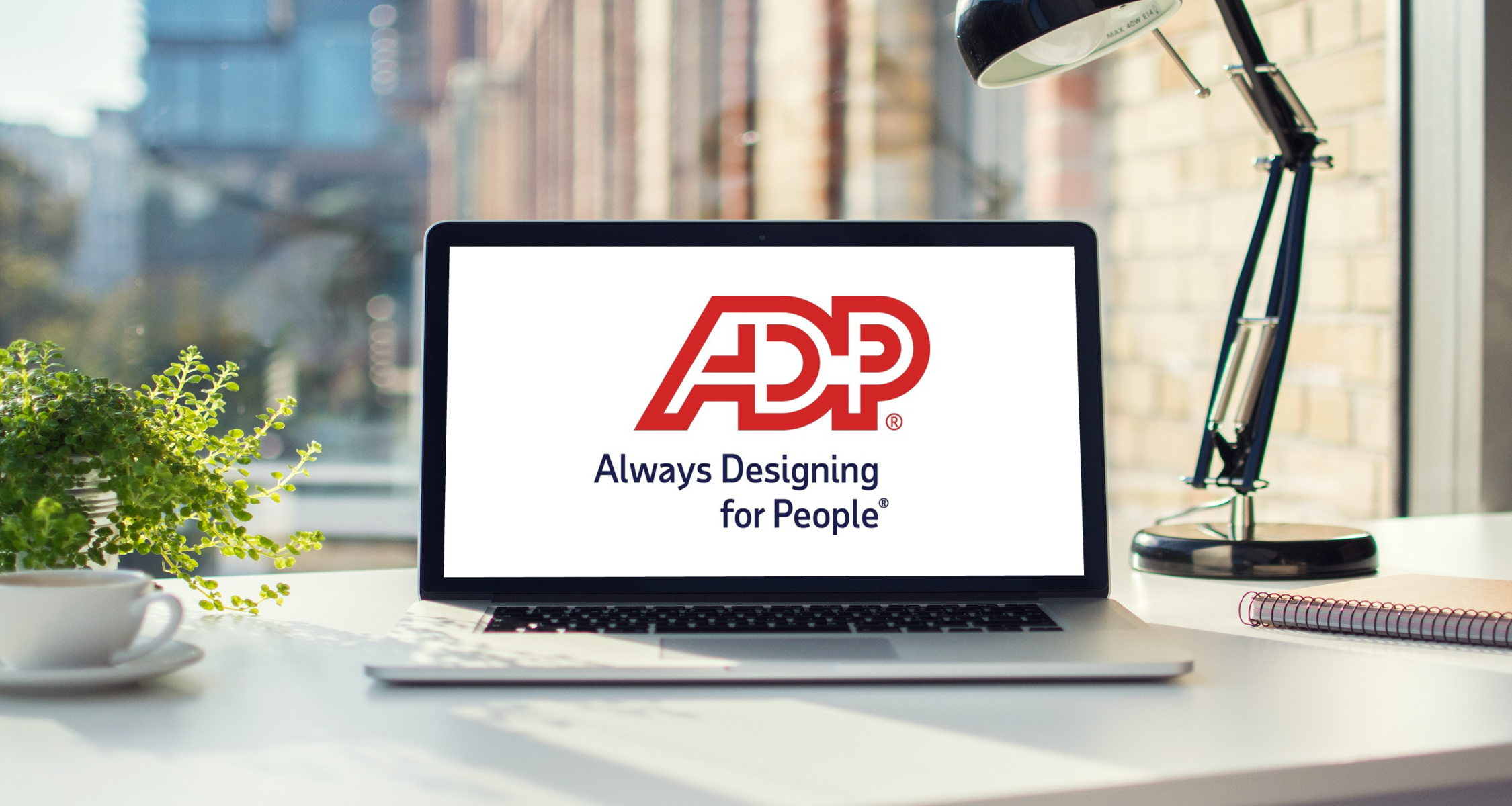 A laptop displaying the ADP logo sits on a desk in front of a sunny window