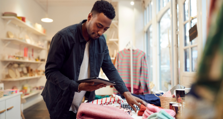 A young Black male small business owner checks his stock of sweaters using a digital tablet