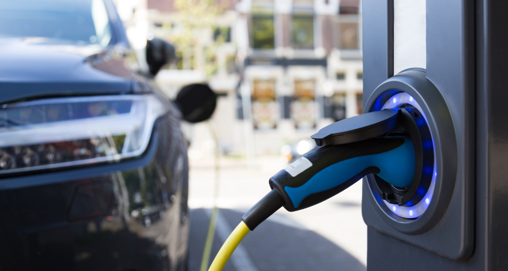 A close-up shot of a glowing electric car charging column, with a car parked in the background.