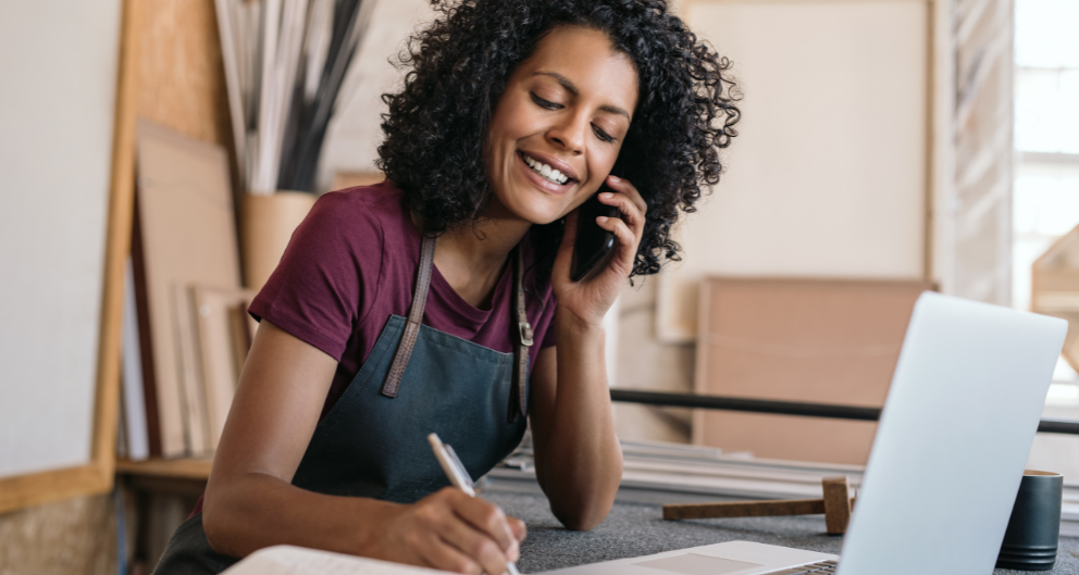 A woman with thick curly hair wearing an apron sits at her desk in front of a laptop. She is talking with a cellphone in one hand and writing in a notebook with the other.