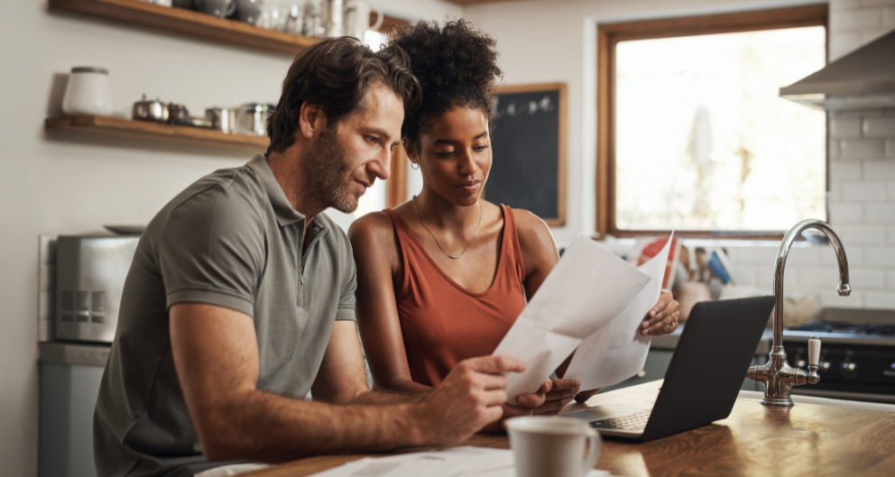 A man and woman sit at their kitchen table together, looking over paperwork in front of a laptop, working on their taxes as married taxpayers