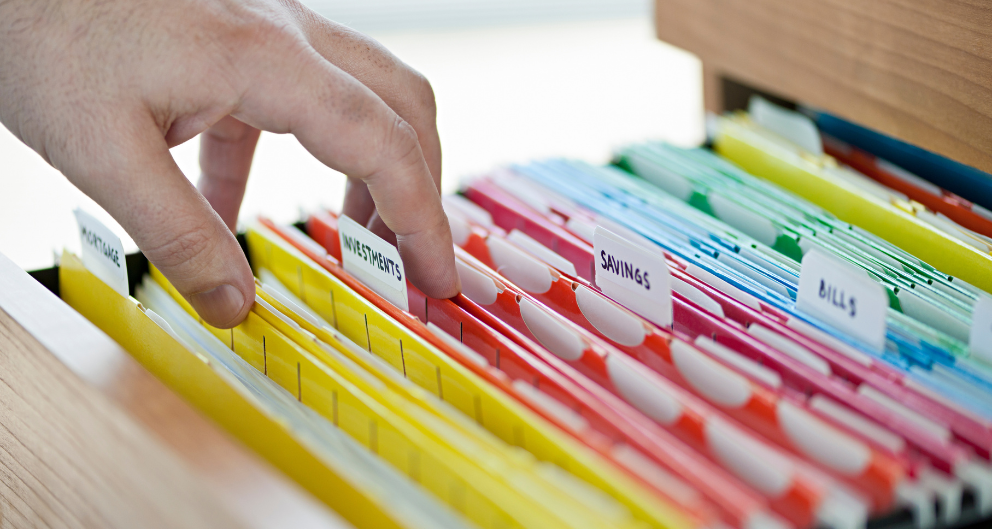 A hand sorts through a drawer full of colorful file folders labeled with names such as bills, investments, taxes, etc.