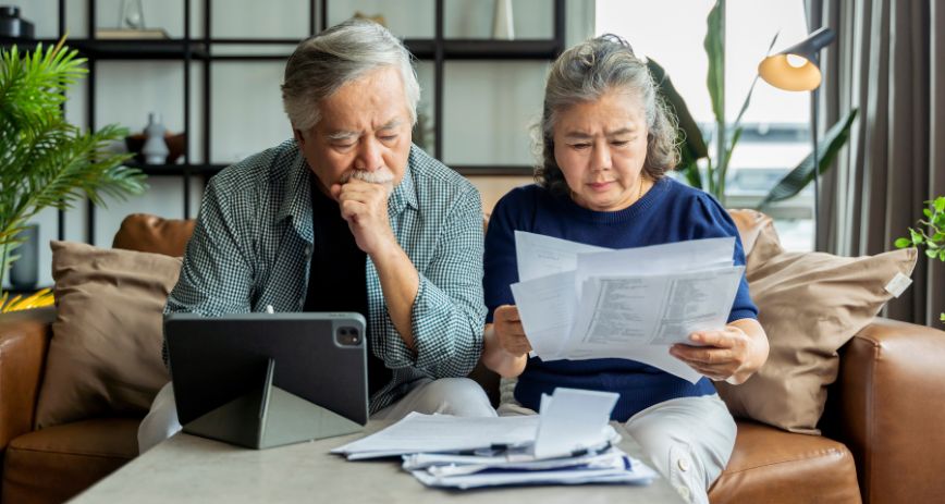 An older Asian couple sit looking over paper work at their computer, with concerned expressions on their faces