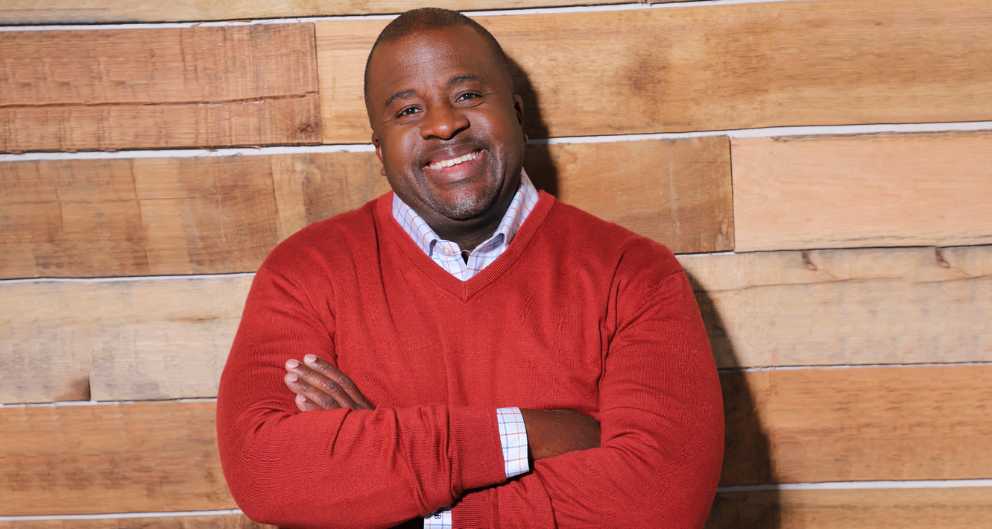 Eugene Brooks, a Black man in a red sweater, leans against a wooden wall. He is the Director of Diversity Marketing for the Atlanta Braves baseball team.