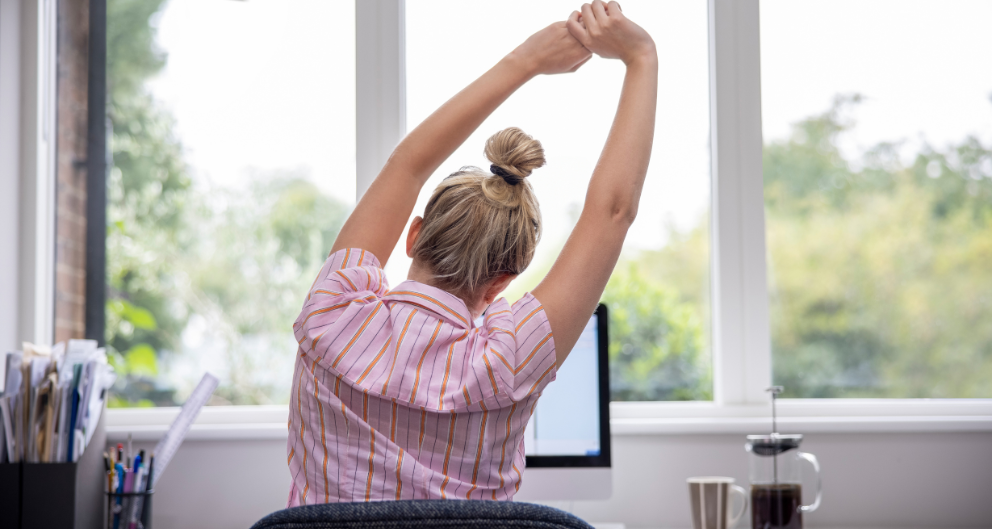 A blonde woman sits at a desk in front of a window, facing away from the camera. She is leaning to the side and stretching her arms high above her head.