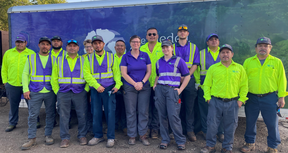 Jessica Riedell stands in the center of a group, wearing a purple shirt. She is surrounded by her landscaping crew, wearing neon green jackets.