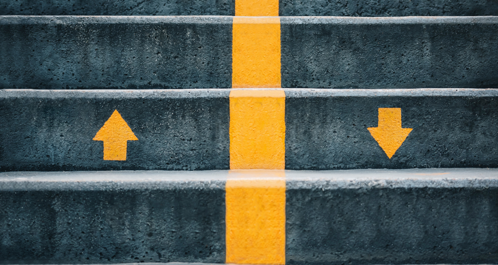 An up close shot of dark grey stairs, with an orange line down the middle and an upward pointing arrow painted in orange on the left, and a matching downward arrow on the right.