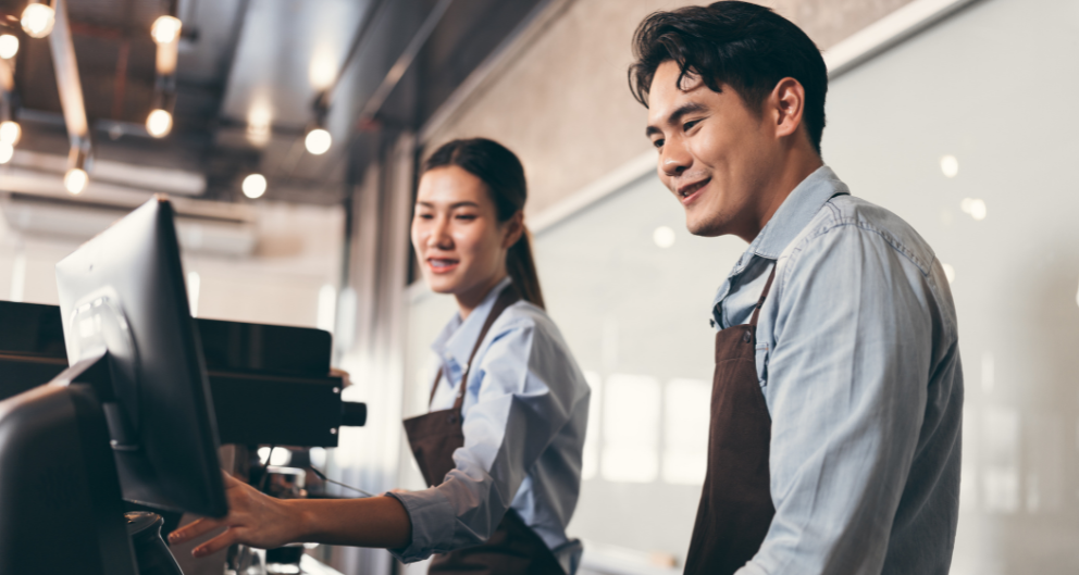 A young Asian man trains a teen girl to work a cash register in a cafe.